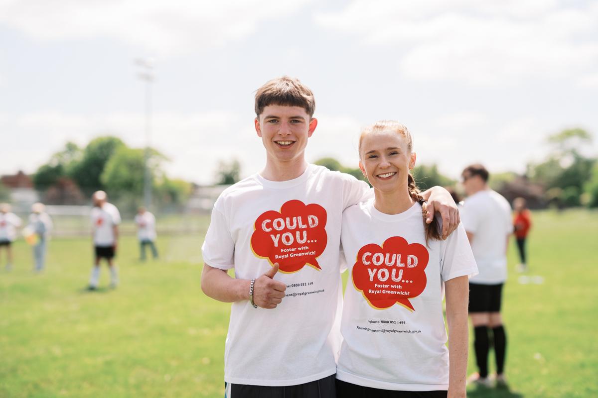 Two people wearing could you foster with the royal borough of greenwich t-shirts - smiling to camera while stood on a football pitch