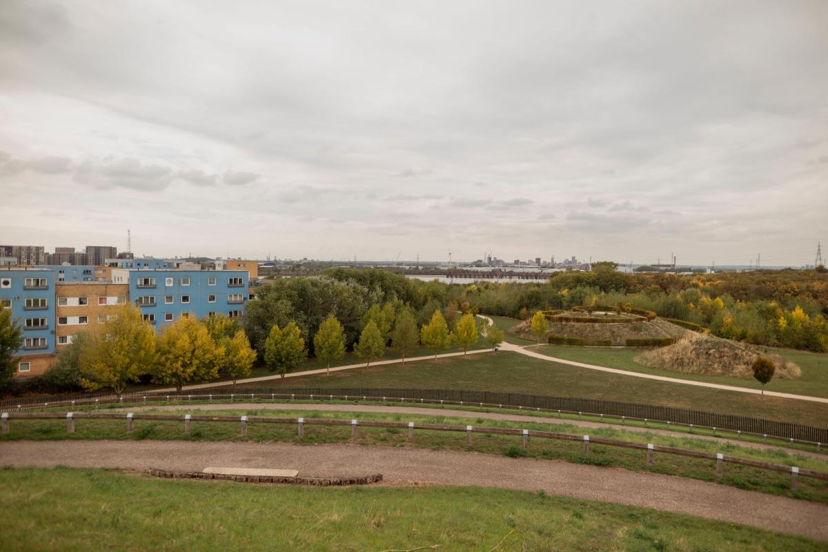 A panoramic shot of Thamesmead Waterfront facing westward, with fields in the foreground, buildings on the left and the riverfront in the background..
