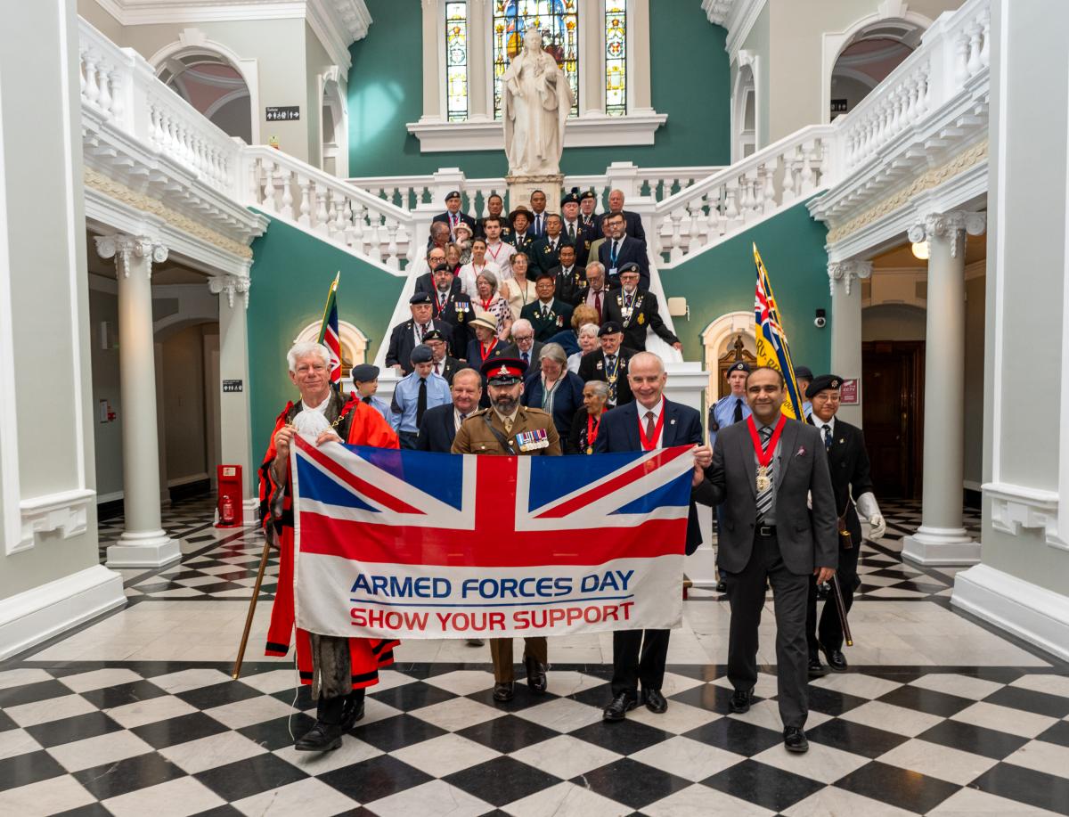 A group of people, some dressed in military uniforms, walk forward through a large hall. The four people at the front are holding a large Union Flag that reads 'Armed Forces Day - Show Your Support'