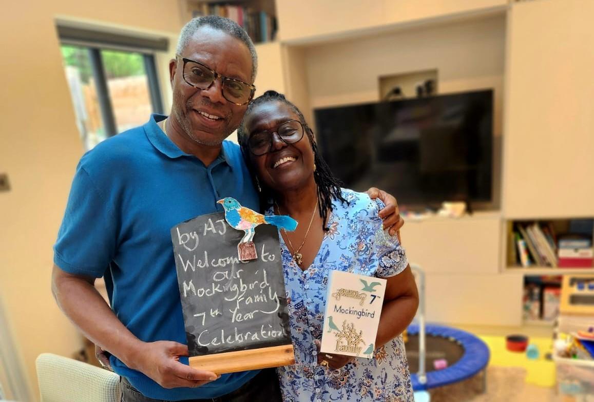 Two people smiling at the camera in a home setting, holding a chalk board and a card marking the anniversary of their mockingbird fostering hub