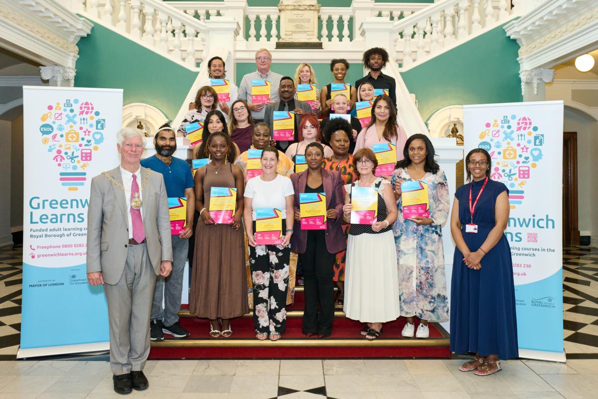 Group of adult learners holding up their certificates on the staircase in the Victoria Hall at the Greenwich Learns awards ceremony, with Cllr David Gardner and Cllr Asli Mohammed standing either side.