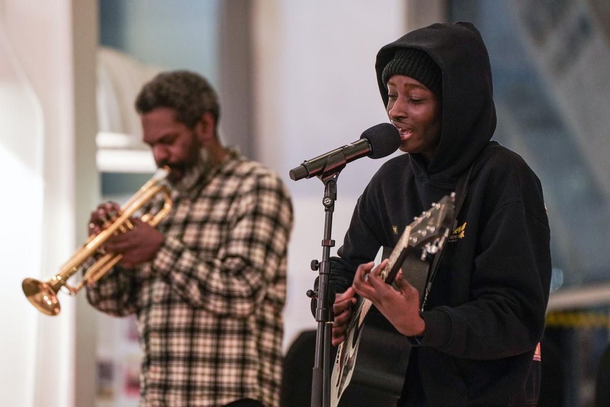 A young person playing guitar and singing. Behind them a person is playing trumpet.