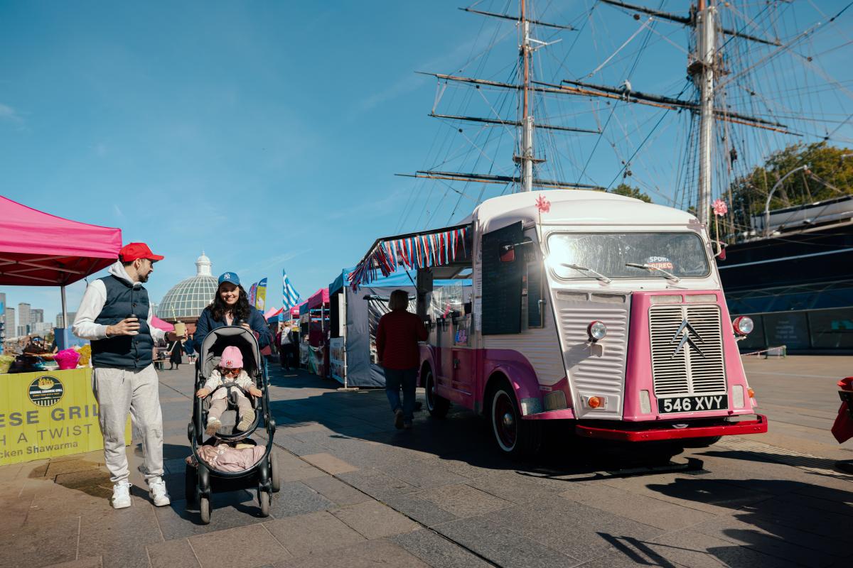 People walking through an outdoor market near the Cutty Sark, with a pink vintage food truck, market stalls, and a stroller in the foreground.