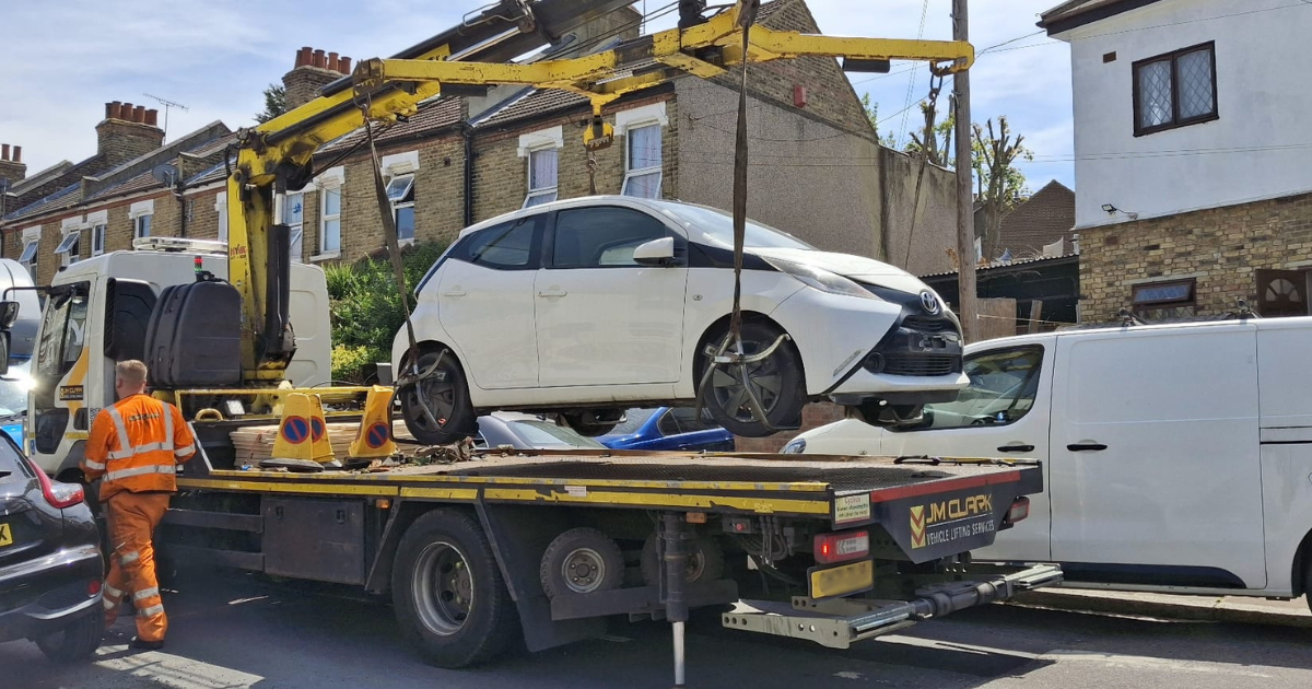 A white car is being lifted onto a recovery truck by a crane on a residential street, with a worker in high-vis standing nearby.