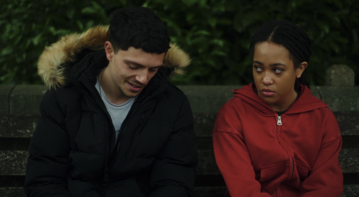 Two people sit on a bench, a man in a hooded jacket and a teenage girl wearing a red hoodie.