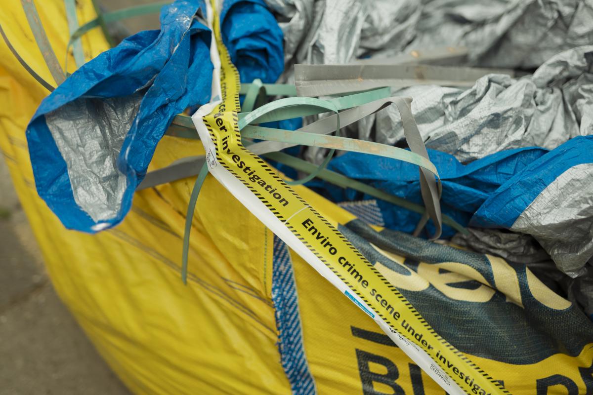 Close-up of a large yellow waste sack filled with blue and grey material and plastic strapping. Yellow environmental enforcement tape reading “Enviro crime scene under investigation” is draped across the waste, highlighting an investigation into illegal dumping.
