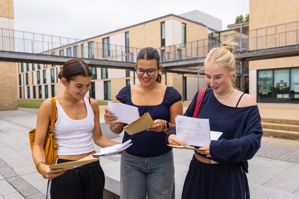 Three young women look at pieces of paper showing their exam results smiling outside a school.