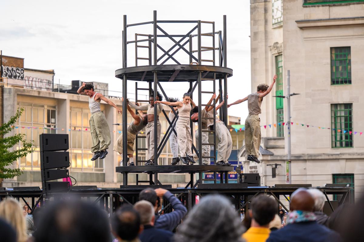 GDIF performers on a raised outdoor stage around a metal structure in General Gordon Square, with an audience watching.