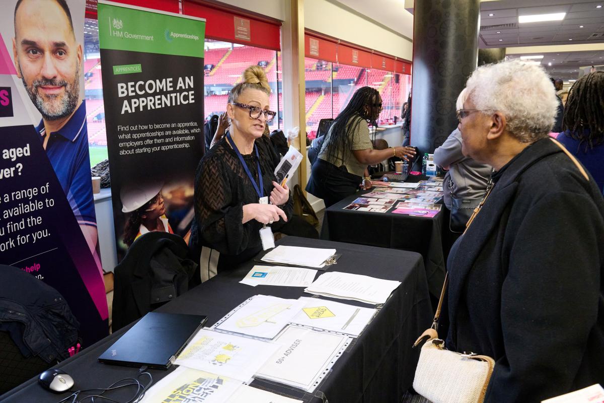 A woman at an employment fair stall speaks to another woman across the table. At the stall is a pull-up banner that reads 'BECOME AN APPRENTICE'. To the right are windows, through which red stadium seating is visible.
