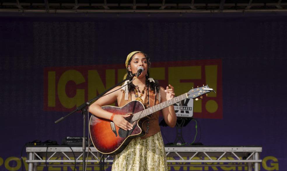 A person on a stage playing guitar and singing. A sign at the back reads Ignite!