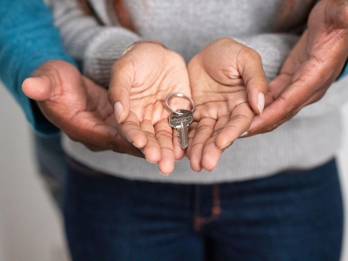 two hands held open with a door key in them and from behind are two hands which is another person supporting their hands gently from underneath