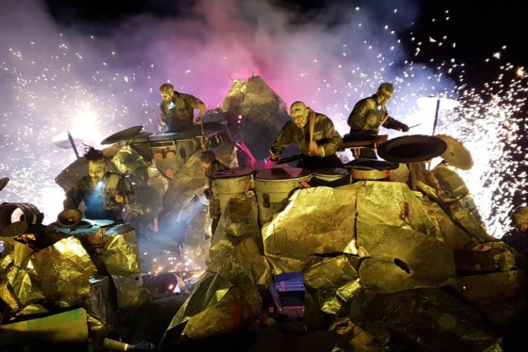 A group of performers dressed in metallic, reflective outfits are seen on a stage that resembles a rocky outcrop. They are engaged in playing various percussion instruments, with dramatic lighting and smoke effects creating a dynamic and intense atmosphere. Bright flashes of light and sparks enhance the visual impact of the performance.