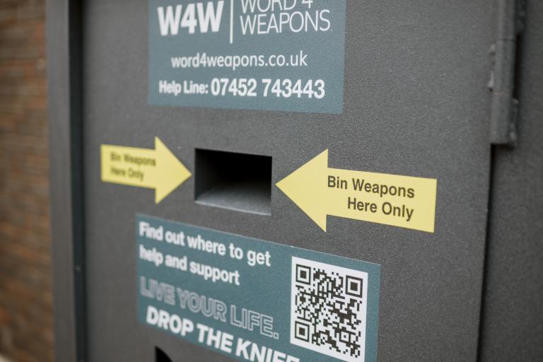 Image of a knife amnesty bin with two yellow arrows pointing to the opening of the bin. Sticker on the bin reading find out where to get help and support. Live your life, drop the knife' and a QR code.