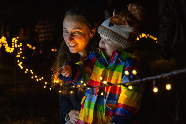 A woman and a young child dressed in a rainbow coloured jacket look at fairy lights