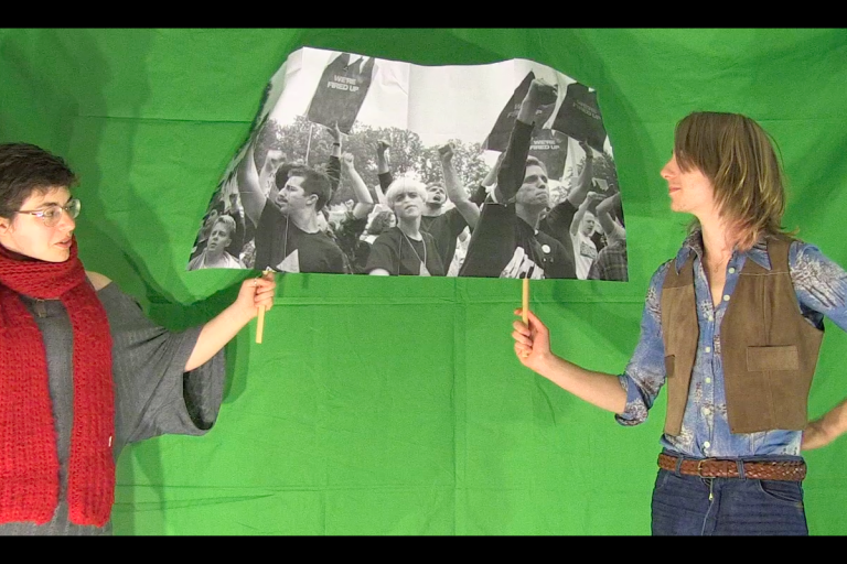 Two people standing facing each other in front of a green screen, holding up a black and white photo of a protest march