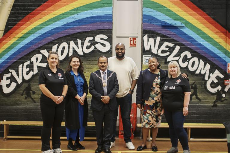 A group photo of youth hub staff and councillors standing in front of a painted mural wall which says 'everyones welcome'