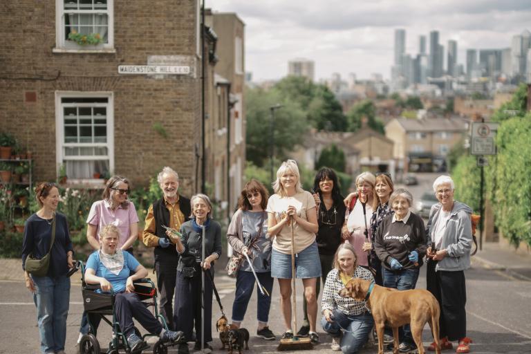 A group of people standing in the road smiling. There are also two sausage dogs and a big brown dog, and the people are holding litter picking and weeding equipment.
