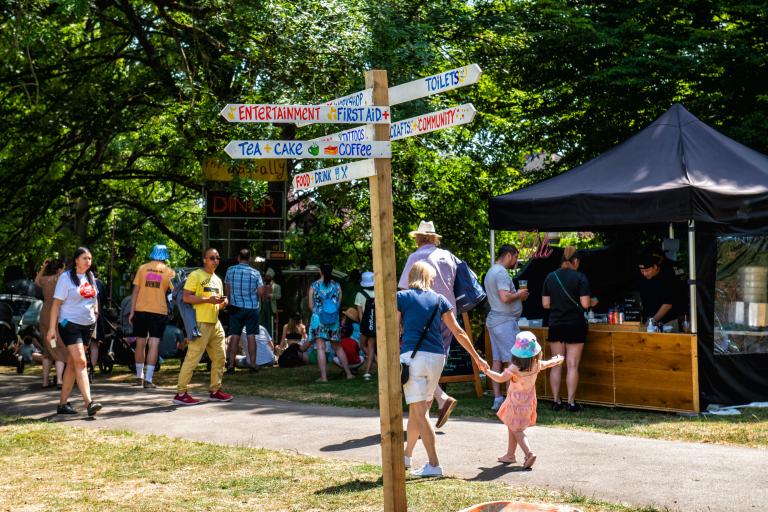 A sign post at an outdoor event, pointing in different directions as people walk past.