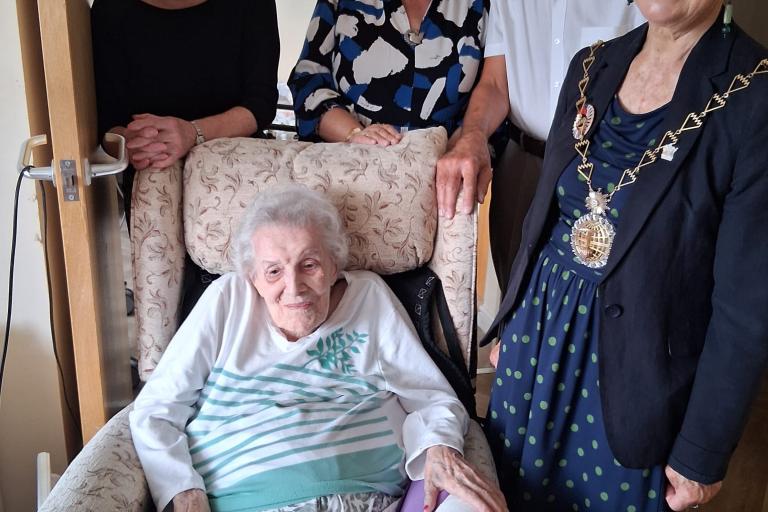 WWII veteran Elsie Vickers is sat in a chair with Cllr Linda Bird standing to her left. Two women and one man are stood behind them. 