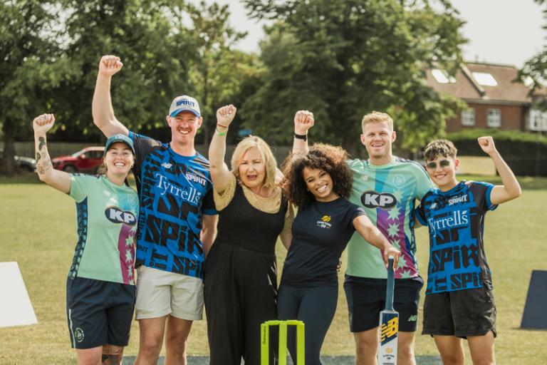 A group of five people standing on a green artificial cricket wicket with their hands up in the air to cheer