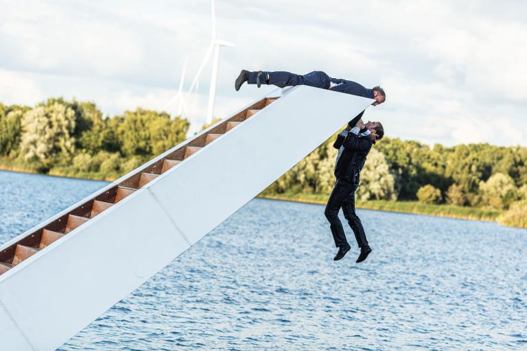 Two people hanging from a white structure floating on a lake, with green trees in the background