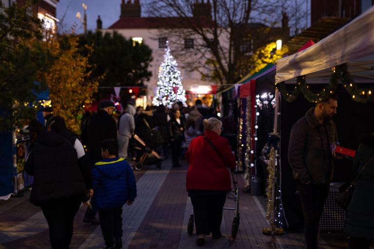 Shoppers browse an evening Christmas market. In the background, there is a lit up Christmas tree.