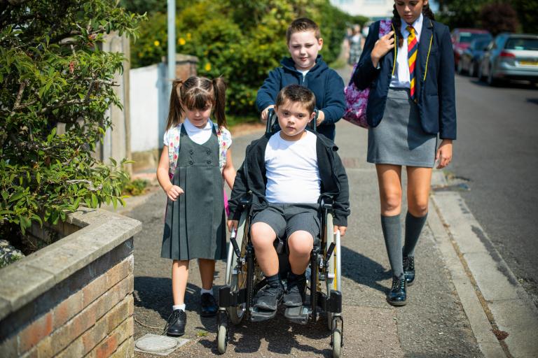 Four young students on their way to school while one of them pushes another on a wheelchair.