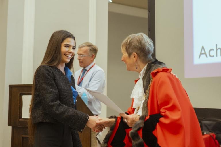 The mayor, in red mayoral gown, shakes hands with a young person who is smiling