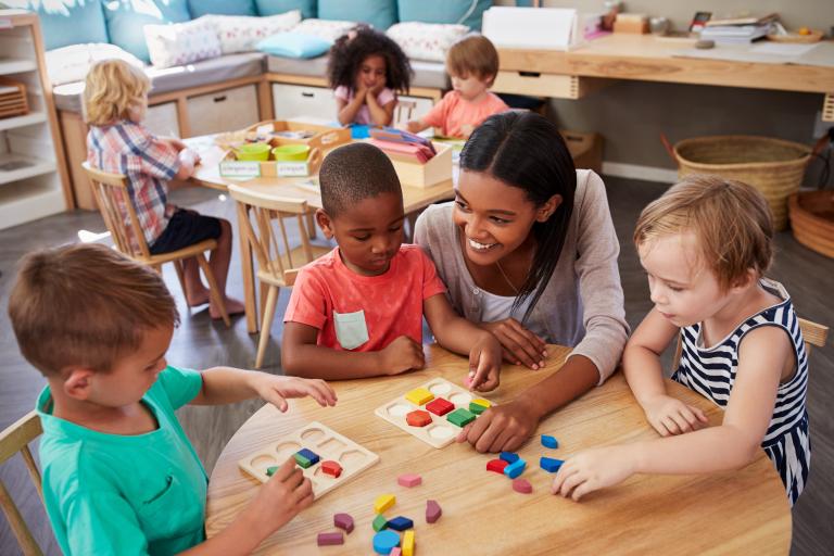 A teacher helping three young children with blocks at a table.