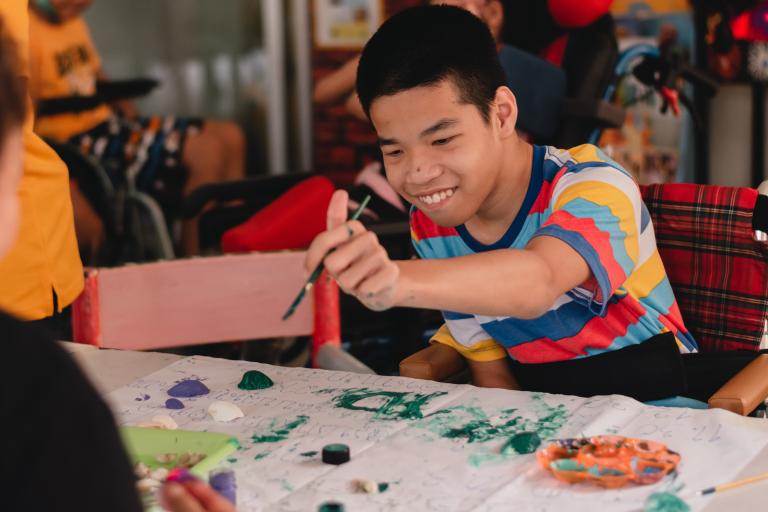 A young boy reaching over the table with a paintbrush.