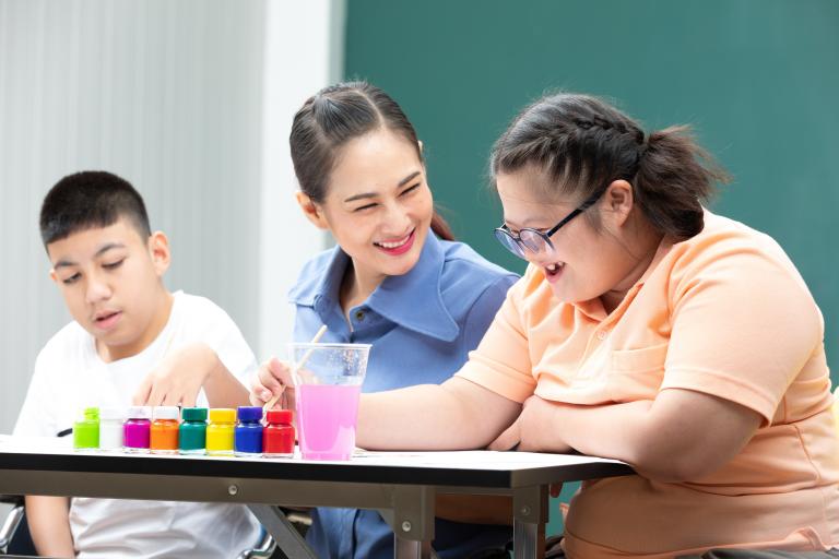 Two students painting at a table while a woman in the middle smiles at one of them.