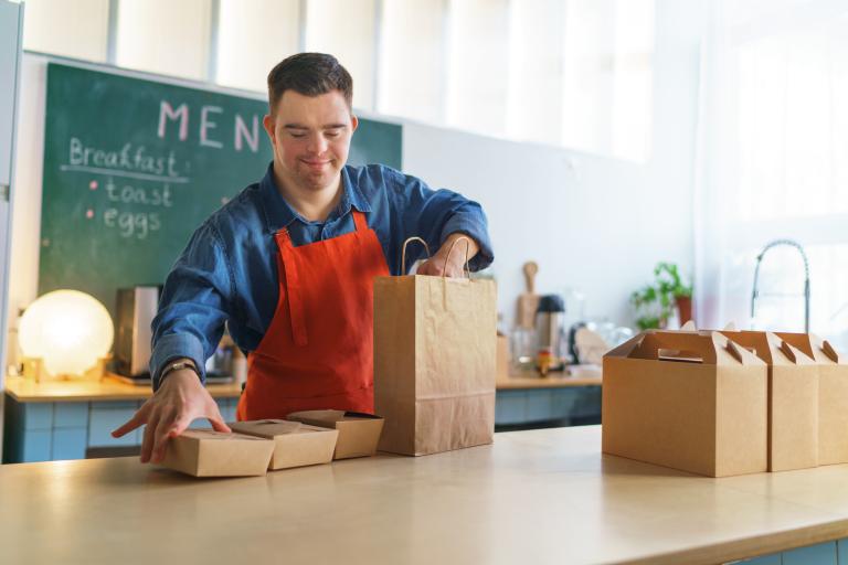A young man in an apron packing brown containers into a paper bag.