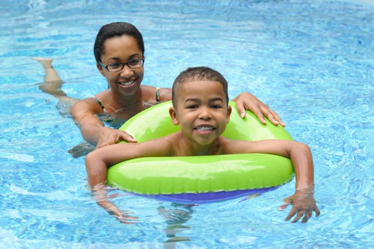 A woman helping to push a young boy in a green floatation ring in the pool.