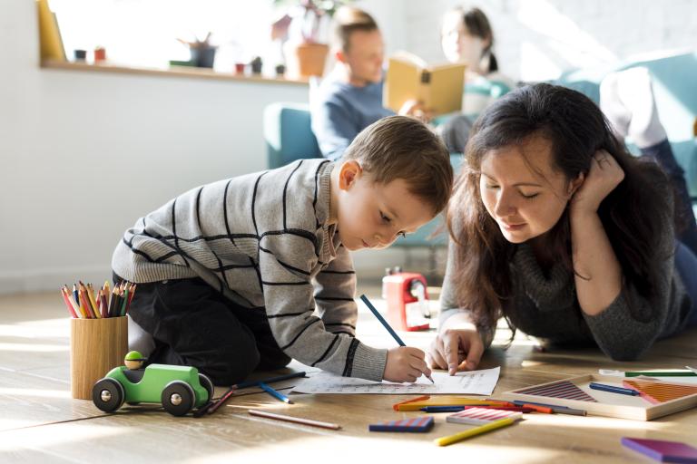 A woman pointing at a piece of paper while a young boy writes with his pencil. In the background, a man is reading to a girl.