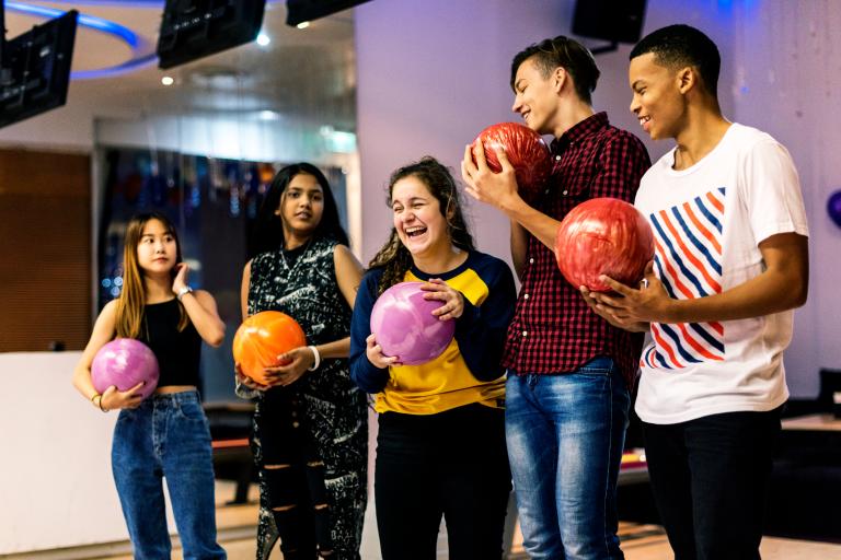 A group of laughing teenagers with bowling balls in their hands at a bowling alley.  
