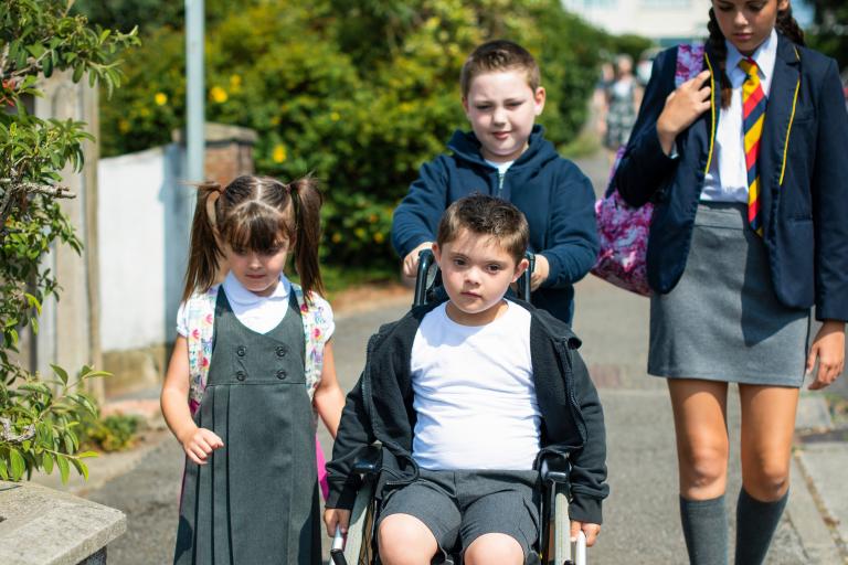 Four young students on their way to school while one of them pushes another on a wheelchair.