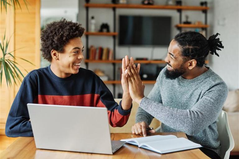 A young man high fiving an older man. One of them has a laptop in front of them while the other is pointing at a book.