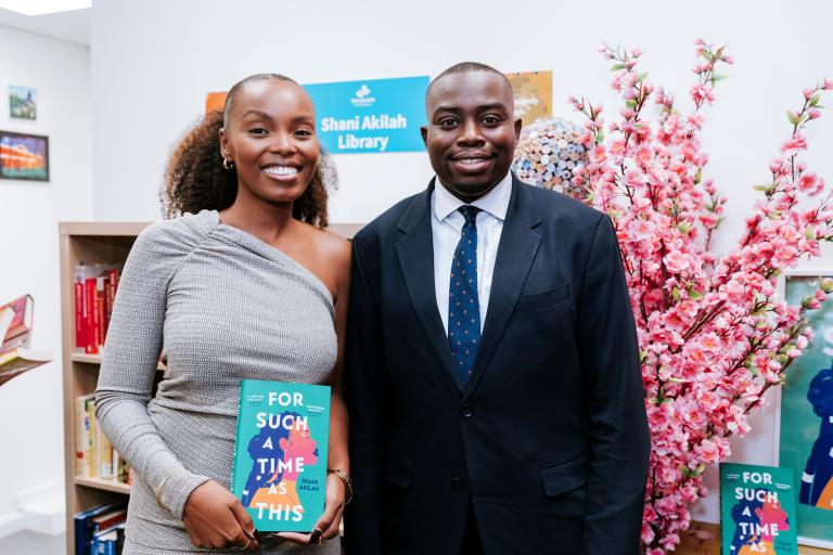 A woman in a grey one shoulder top stands smiling holding a book. To her left is a man in a suit smiling. They are both looking to camera. Behind them is a pink cherry blossom display, a bookcase and a sign that reads: The Shani Akilah Library