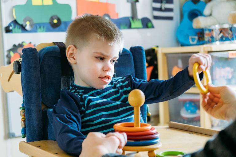 A young boy is propped up at a table and grabbing a plastic ring that someone is holding up to him.