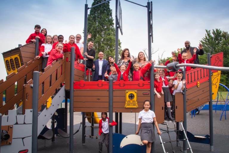 A group of schoolchildren in uniform with a few well-dressed adults in the middle clustered on a brand new pirate ship playground.