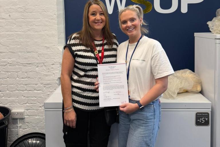 A photo of two people holding the Women's Safety Charter in front of a board which says 'Welcome to WSUP'