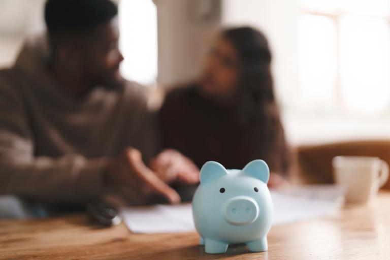 A blue piggy bank on the table while a couple argues over a pile of papers in the background.