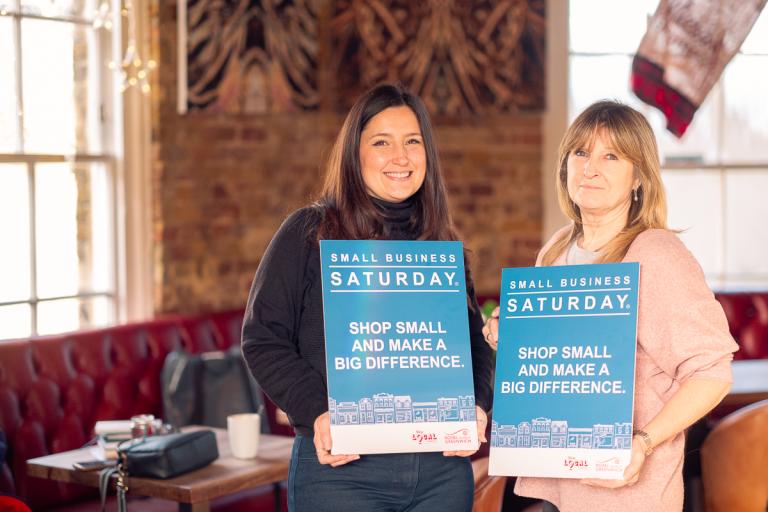 Small Business Saturday - two businesswomen stood smiling to camera, holding Small Business Saturday signs