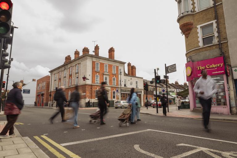People crossing a road at a pedestrian crossing in a busy Eltham Town Centre with shops and traffic lights.