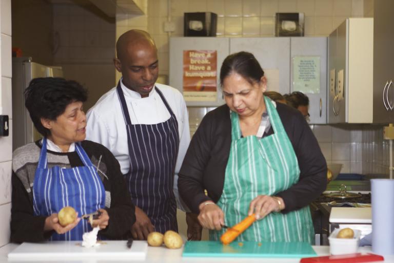 A chef watching two women peel potatoes and carrots in a kitchen.