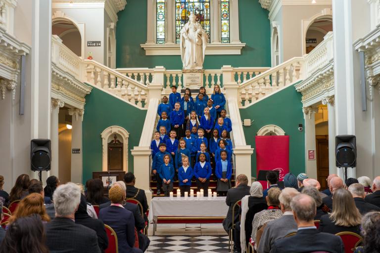 A choir of children wearing blue uniform singing on a large white staircase. In front of them is an audience of people with their backs facing the camera.