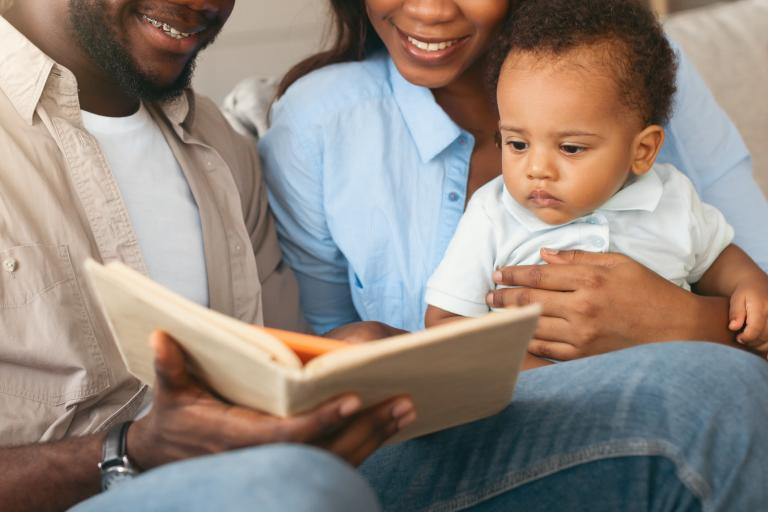 A baby sitting in their mother's lap as their father reads to them from an open book.