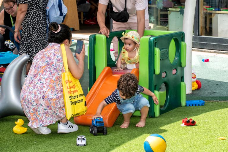A mother taking picture of her toddler on a small slide. 