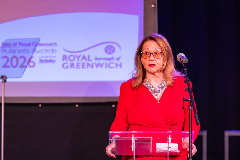 A woman in a red dress and sparkly necklace speaks on stage into a microphone. Behind her is a screen that says Best of Royal Greenwich Business Awards 2026 and the Royal Greenwich logo