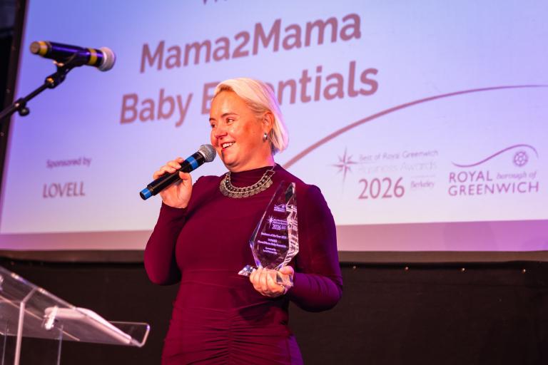 A woman holds a glass trophy in one hand and a microphone in another. Behind her, the screen reads: Mama2Mama Baby Essentials